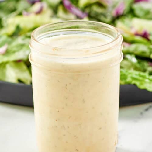 Creamy Italian dressing in a glass jar and a salad behind it.