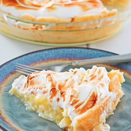 A meringue topped lemon pie slice on a plate and the pie behind it.