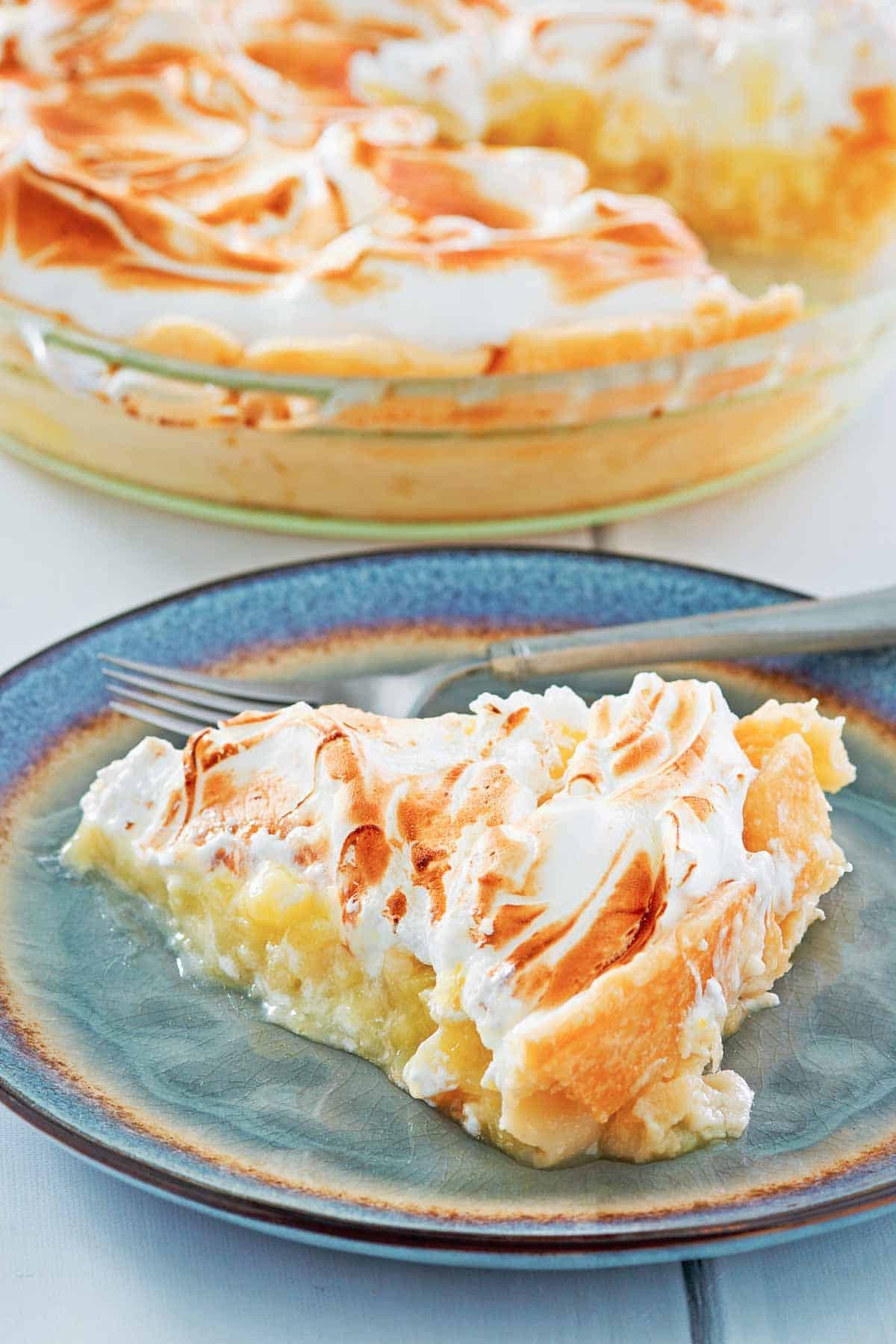 A meringue topped lemon pie slice on a plate and the pie behind it.