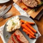 Meatloaf with ketchup glaze, carrots, and mashed potatoes on a plate.
