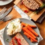 Sliced meatloaf with ketchup glaze on a plate with mashed potatoes and carrots.