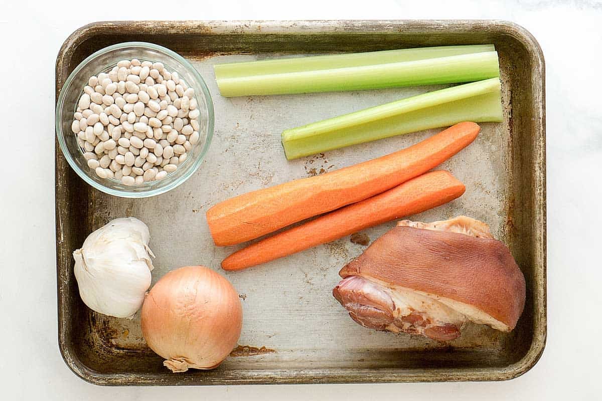 Navy beans, ham hock, celery, carrots, garlic, and onion on a tray.