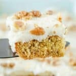 A slice of banana cake with cream cheese frosting on a spatula held over the cake.