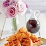 Copycat Grand Lux Cafe chicken and waffles, a small pitcher of syrup, and flowers in a vase.