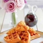 Copycat Grand Lux Cafe chicken and waffles on a plate, a pitcher of syrup, and flowers in a vase.